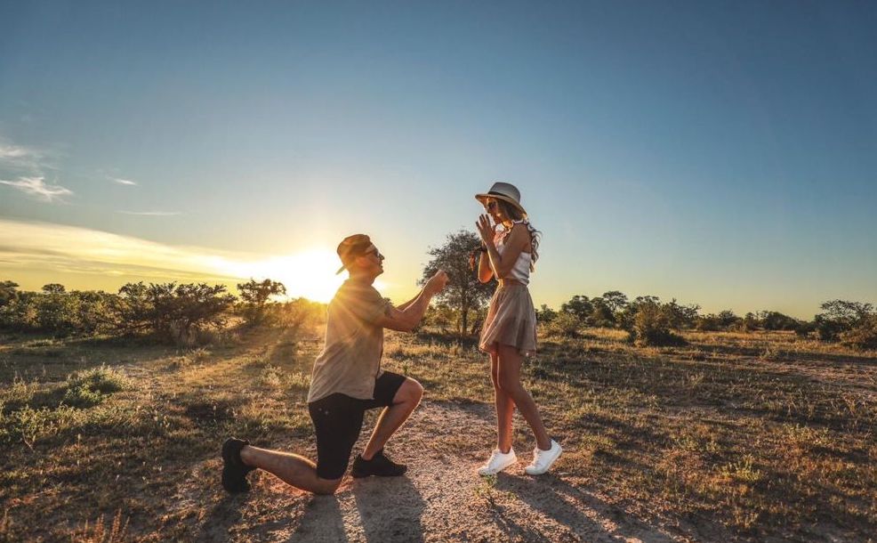 Person kneeling and proposing to another on a rocky path at sunset in a field