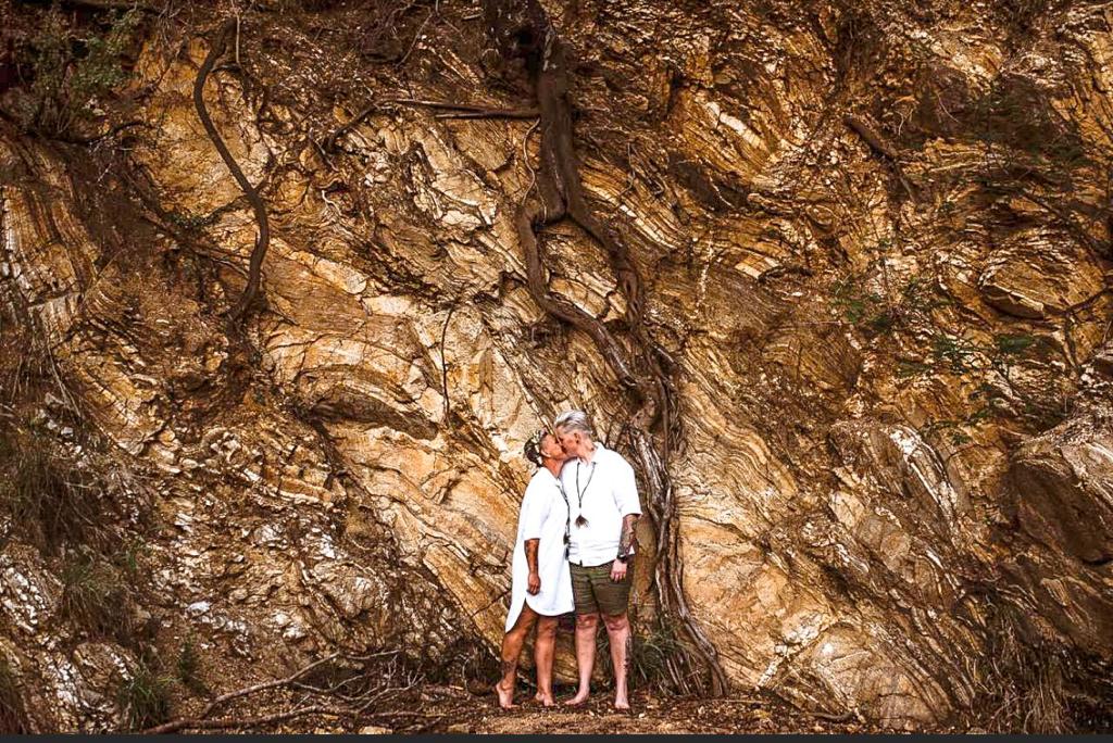 Two people standing in front of a large rocky cave wall, one wearing white and the other shirtless.