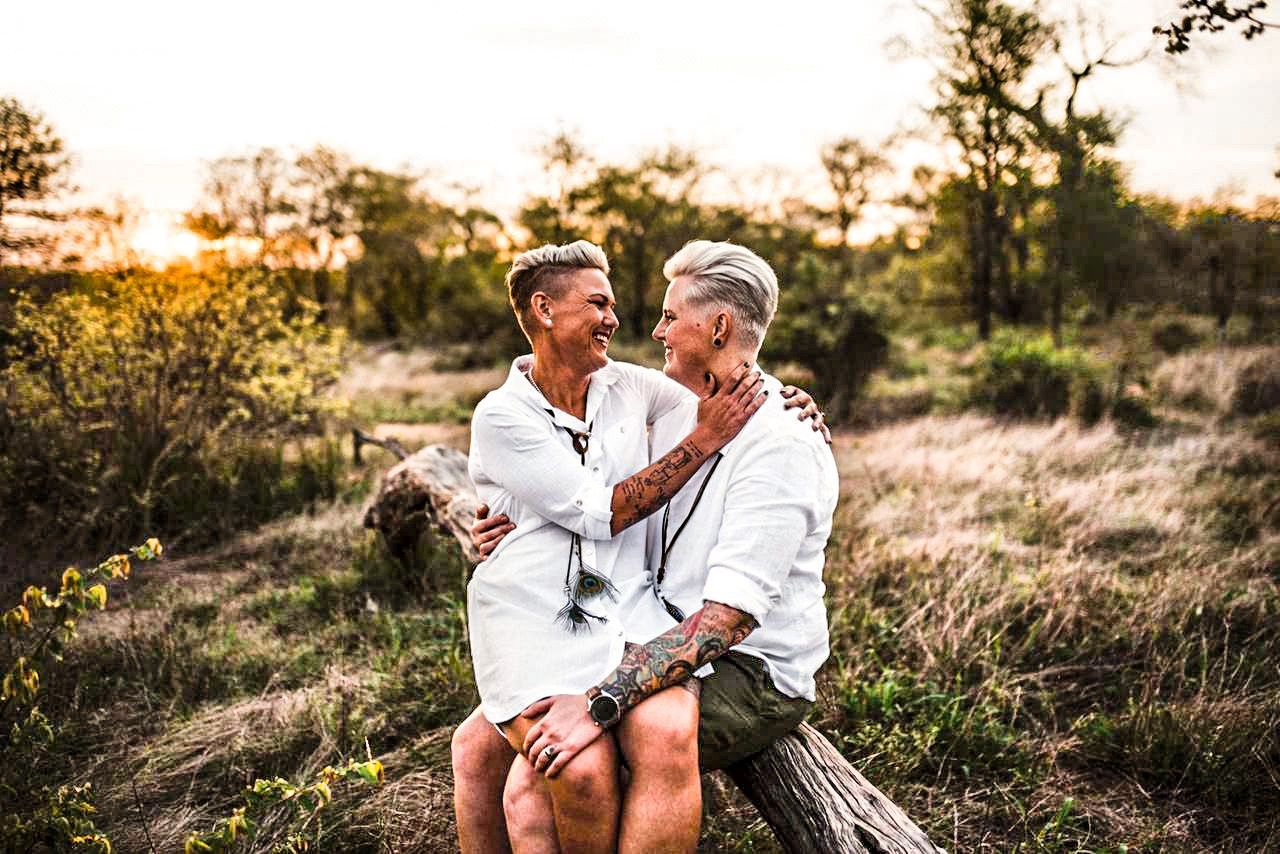 Two people in white shirts sitting on a log in a sunny field, smiling at each other.