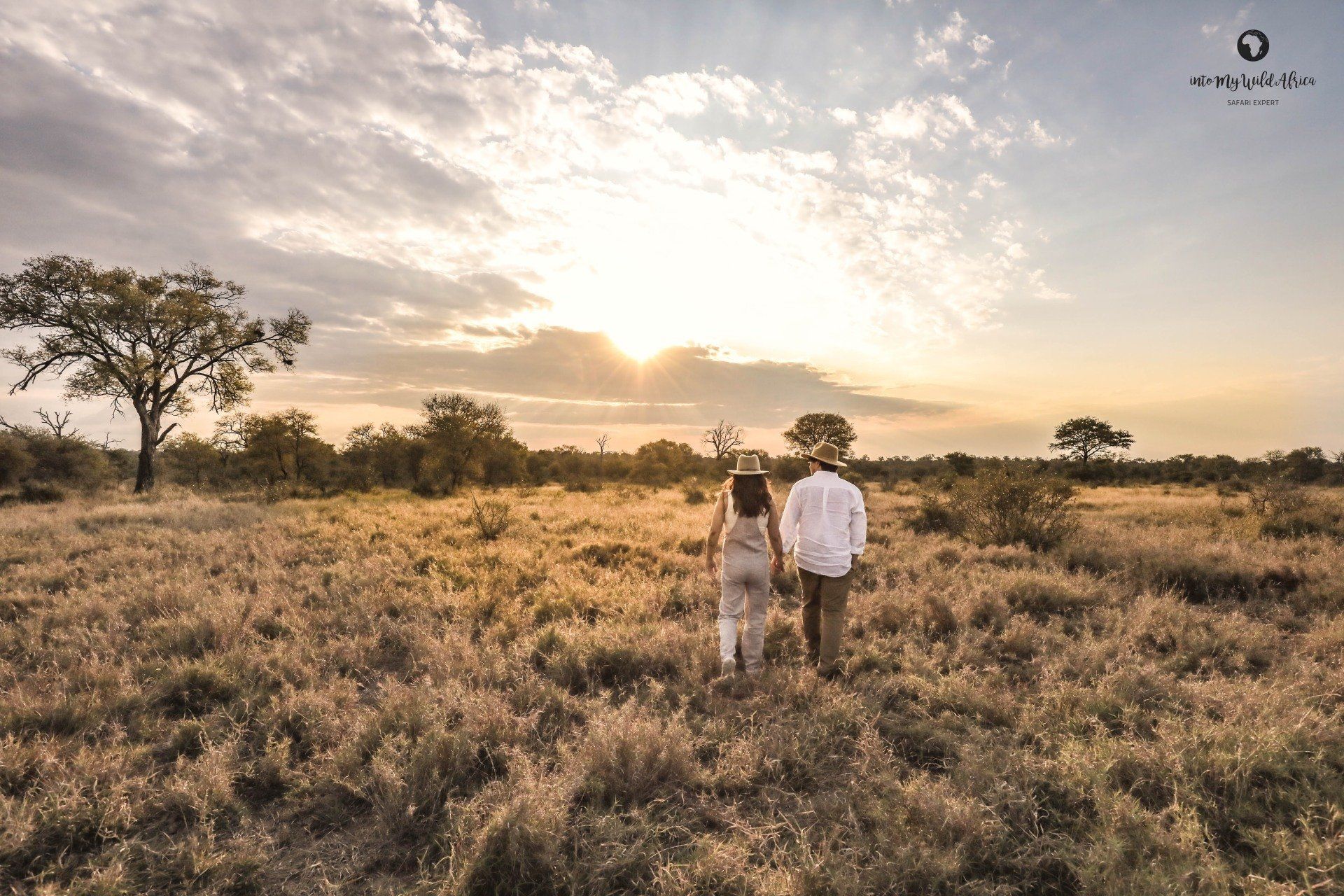 Two people walking through a grassy field at sunset, with scattered trees and warm golden light.