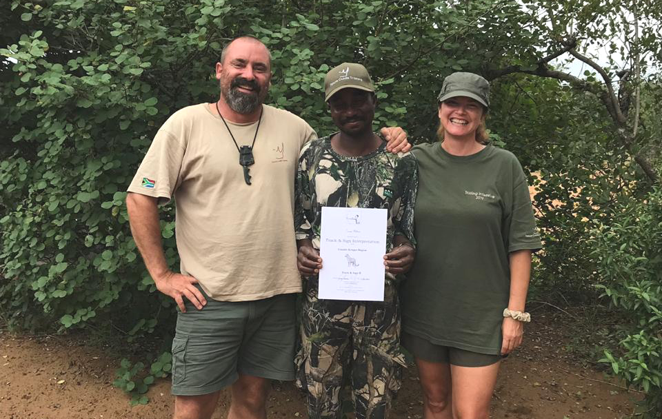 Three people standing outdoors, one holding a certificate, with dense green trees behind them.