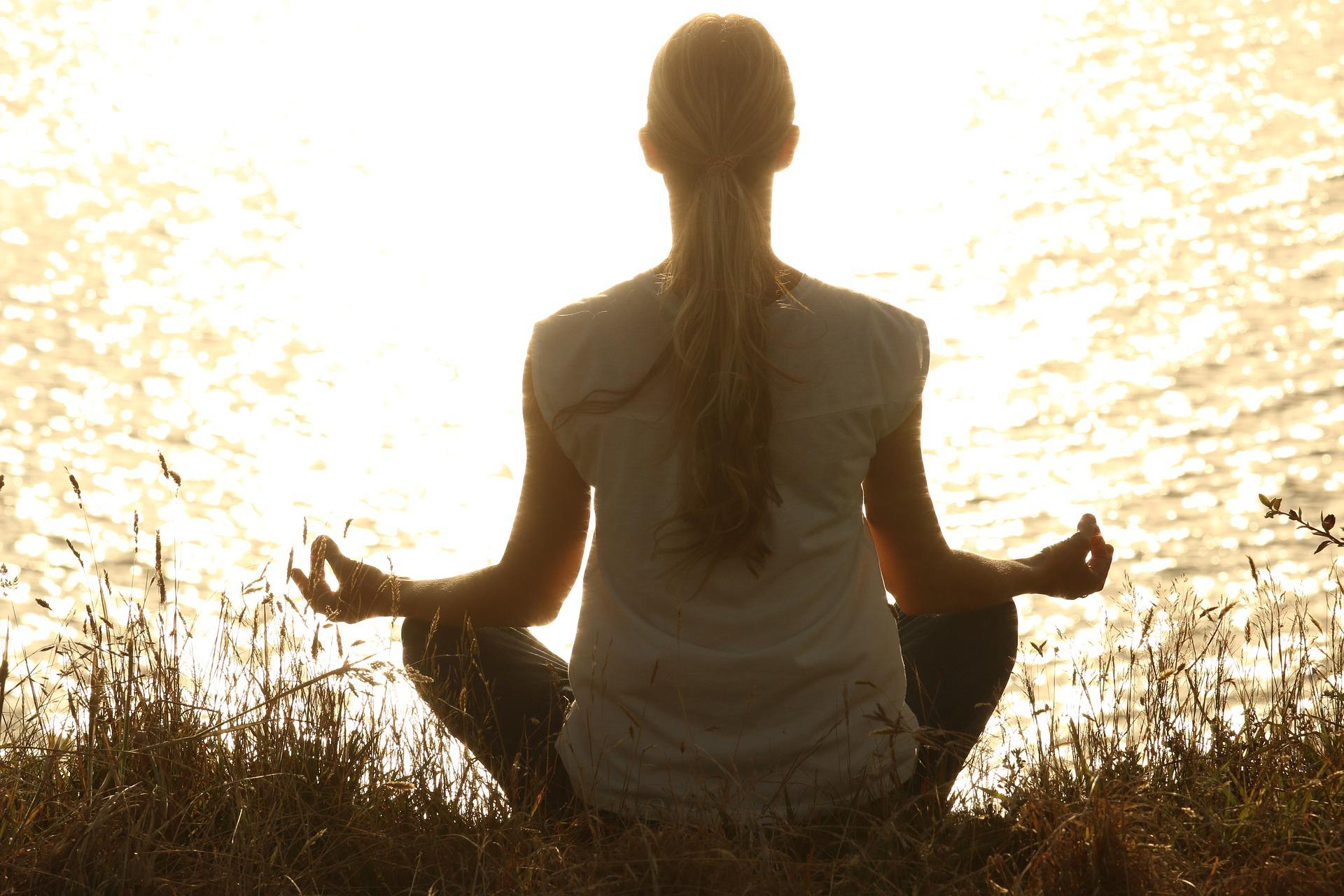 Person meditating cross-legged by a sunlit body of water, seen from behind.