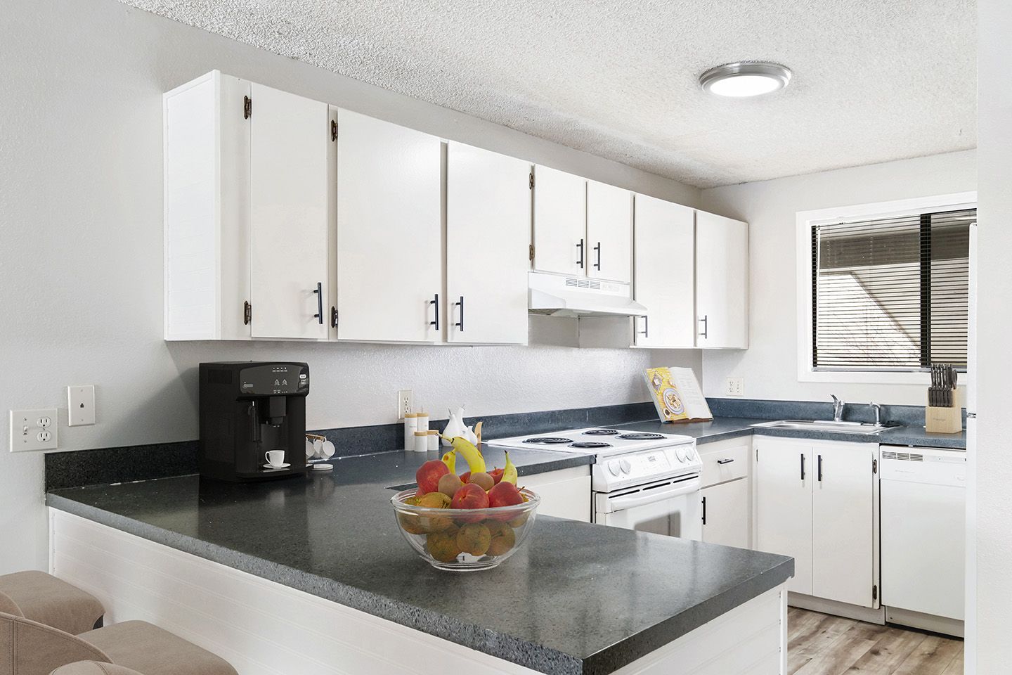 Bright kitchen with white cabinets, dark countertops, a fruit bowl, and a window over the sink.