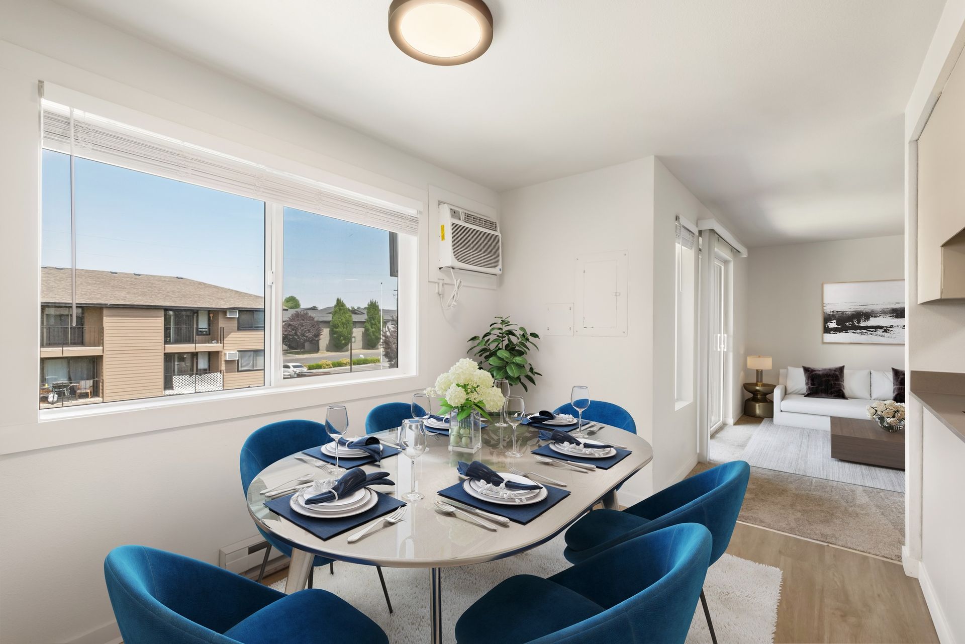 Bright dining area with blue chairs, set table, large window, and a view into a living room