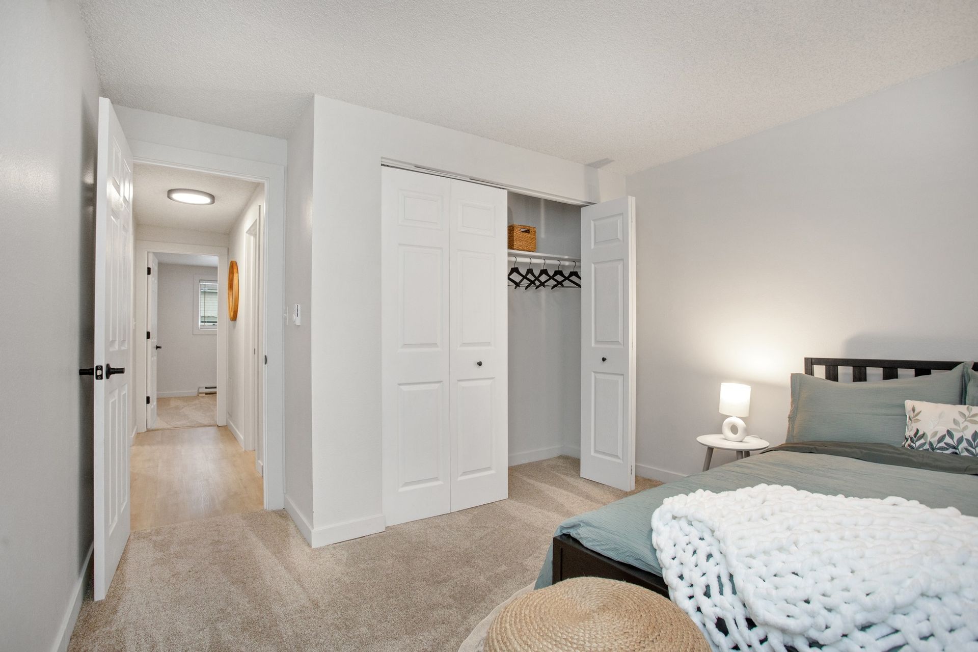 Bright bedroom with open closet doors, hallway view, and a bed with teal bedding and a lit nightstand lamp.