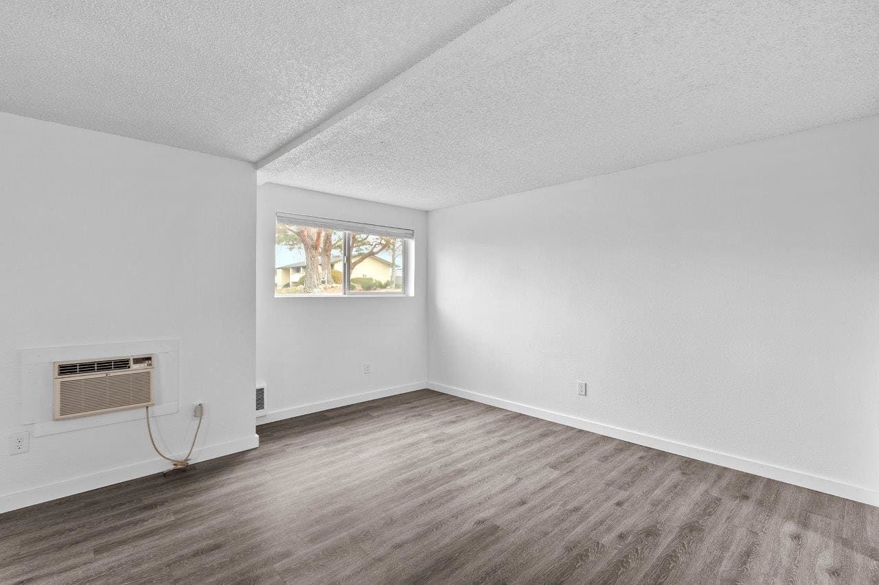 Empty white room with gray wood flooring, a small window, and a wall-mounted heater.