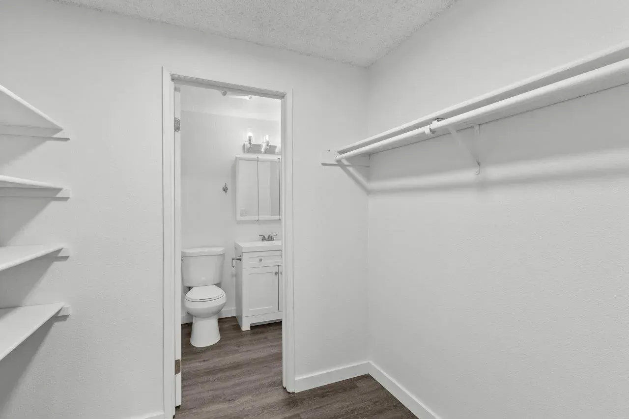 Empty walk-in closet with white shelves and hanging rods, opening to a small bathroom with toilet and vanity.