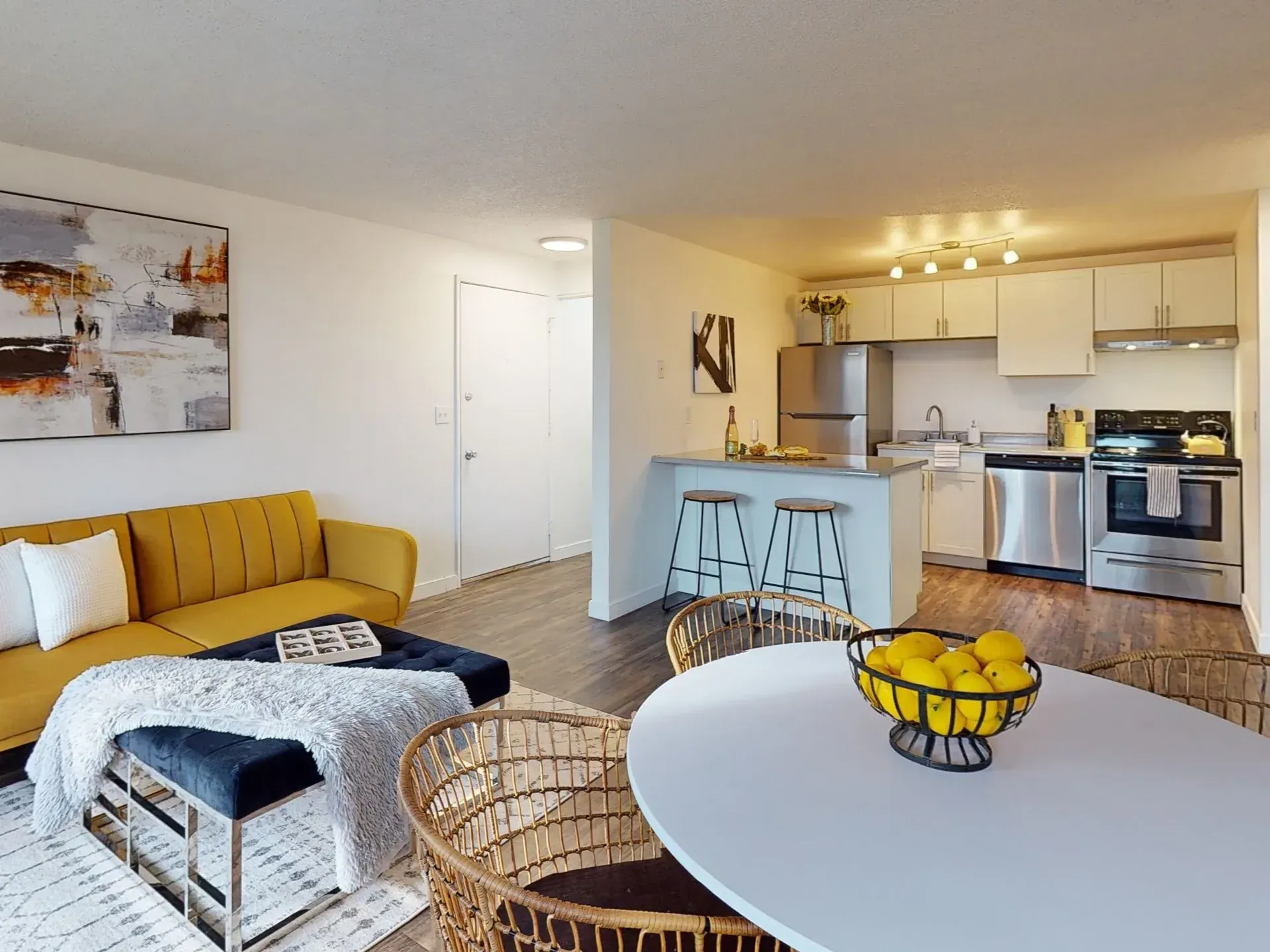 Bright living room with yellow sofa, wicker chairs, dining table, and open kitchen with bar stools
