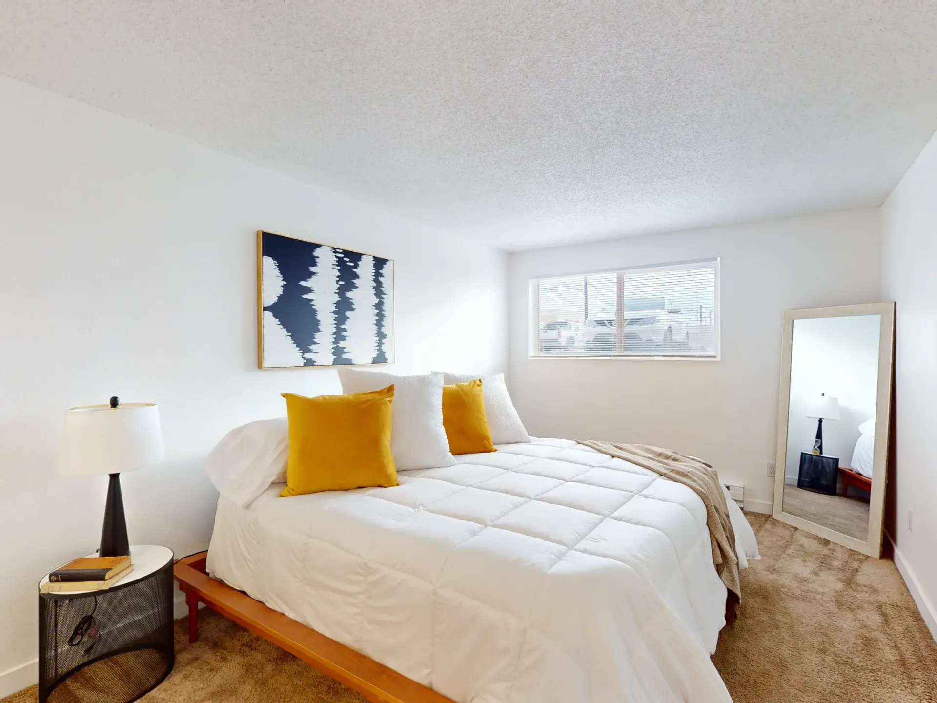 Bright bedroom with white bedding, mustard pillows, wall art, side table lamp, window, and standing mirror