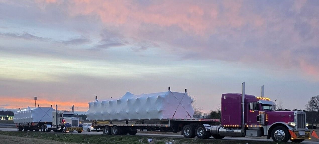 Two semi-trucks transporting large, covered objects on a highway at dusk under a colorful sky.