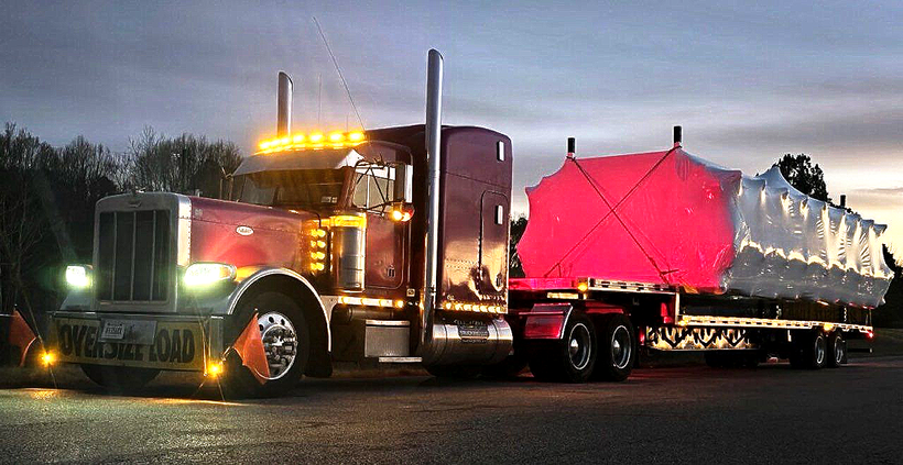 Purple Peterbilt semi-truck with a flatbed trailer loaded with concrete blocks parked outside a warehouse on a sunny day.