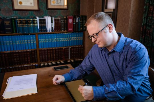 A man in a blue shirt and glasses is sitting at a desk in front of a bookshelf.