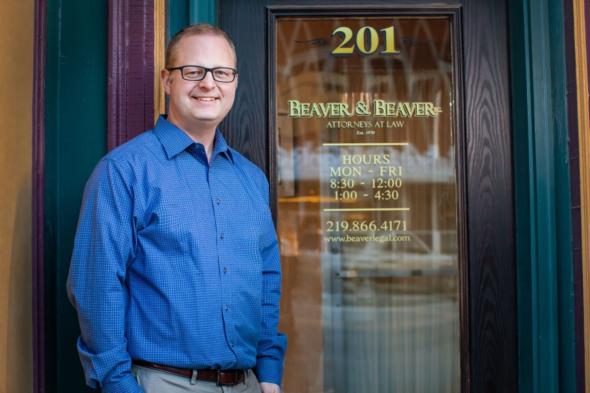 A man in a blue shirt and glasses is standing in front of a glass door.