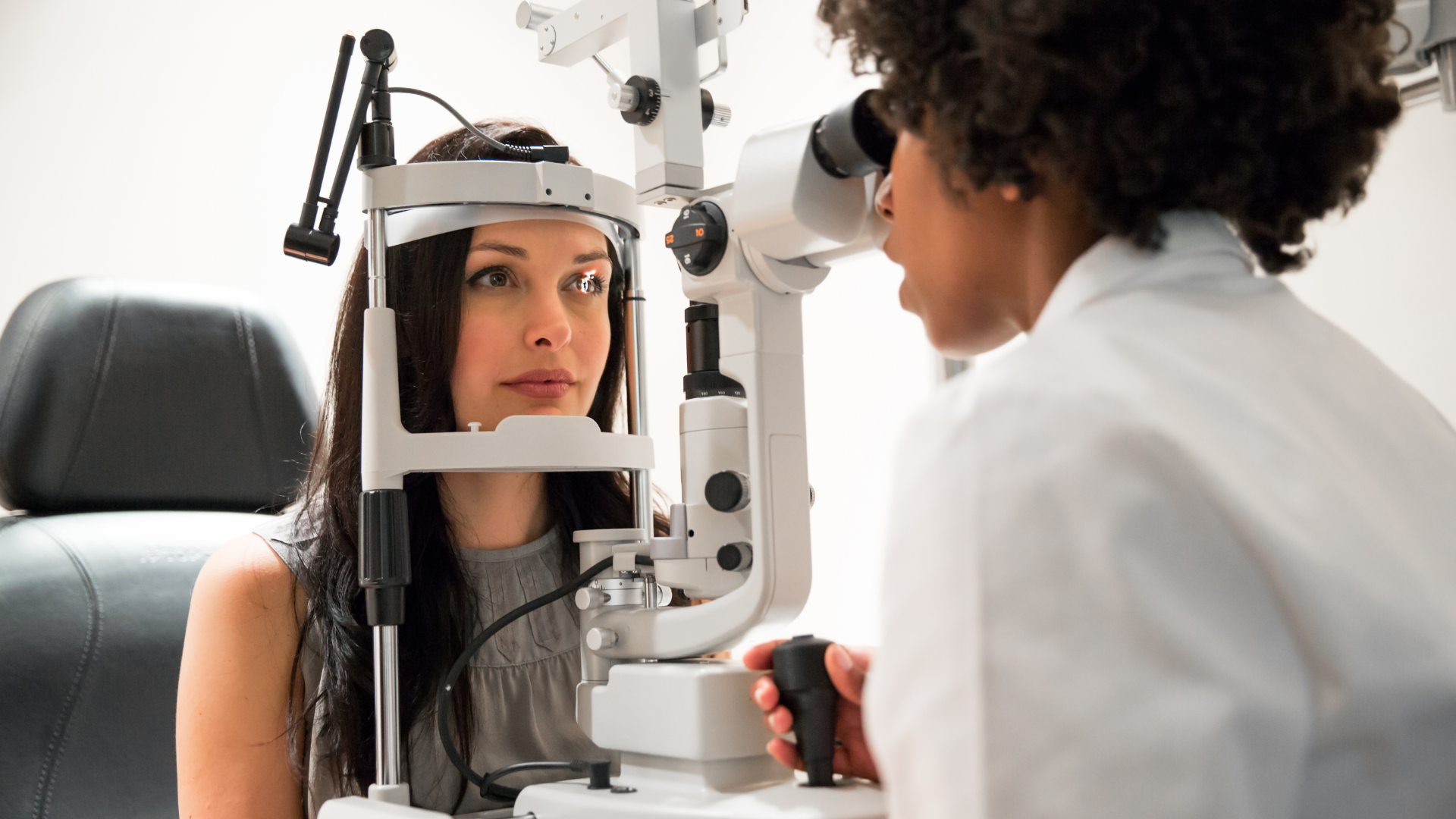 Woman having eye exam with optician using a slit lamp in a white exam room.