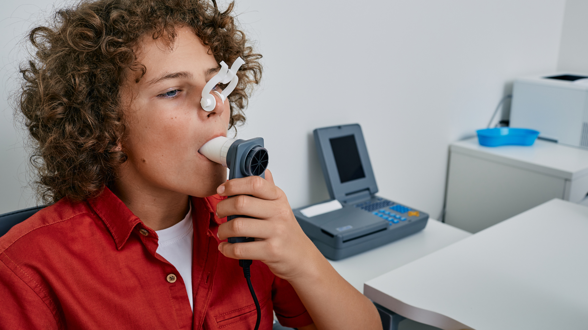Boy with curly hair taking a breathing test in a medical office, exhaling into a device.