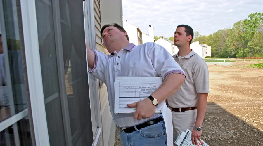 Two men are standing outside of a house looking at a window.