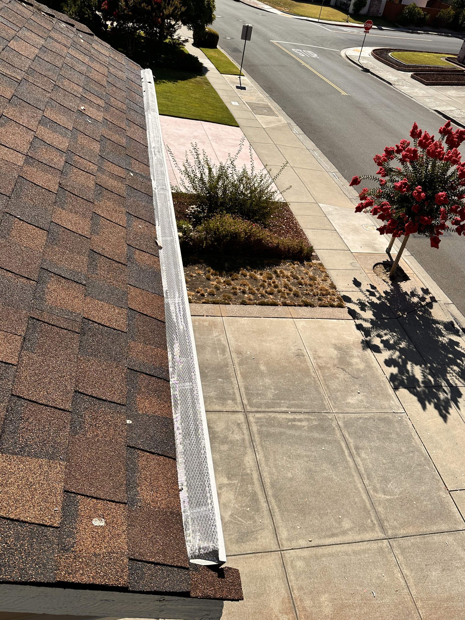 High-angle view of an asphalt shingle roof edge with a metal gutter guard, overlooking a driveway and street.