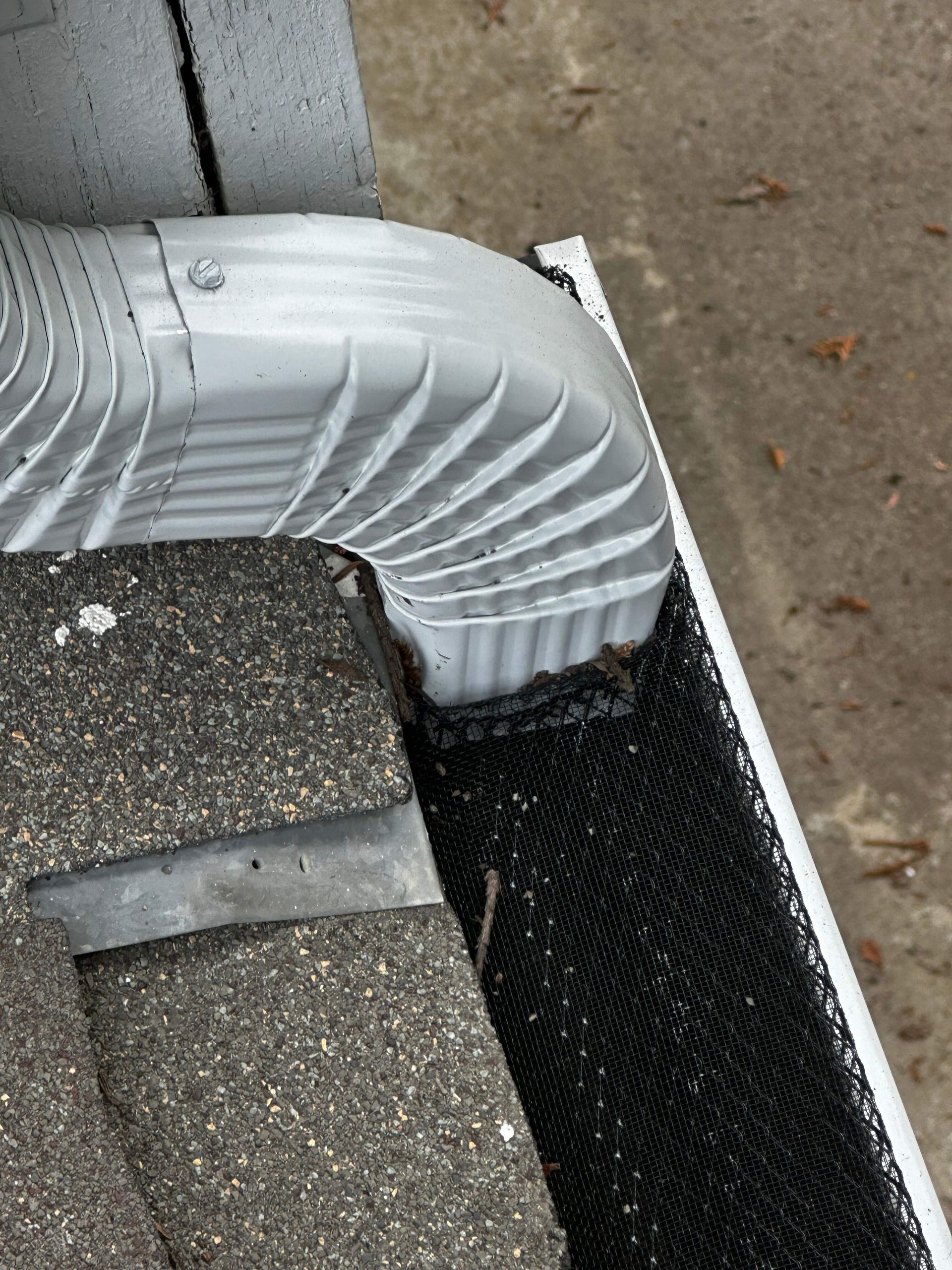 A white corrugated downspout elbow connects to a black mesh gutter guard on a shingled roof edge.