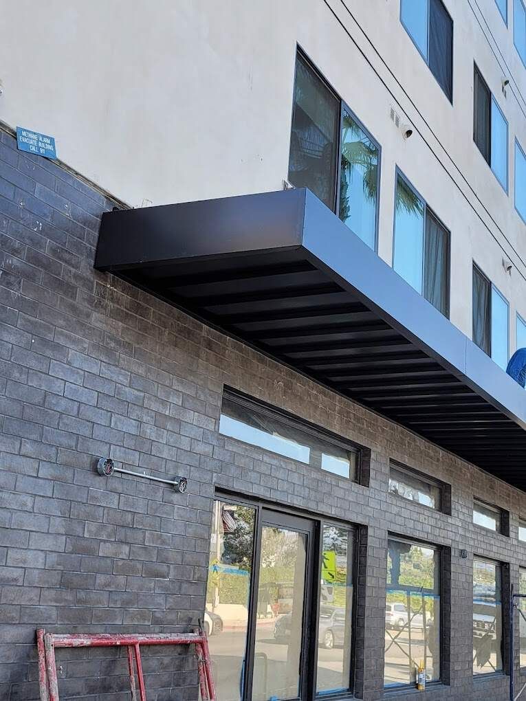 Low-angle view of a modern building entrance with dark brick, glass doors, and a black metal awning under a light wall.