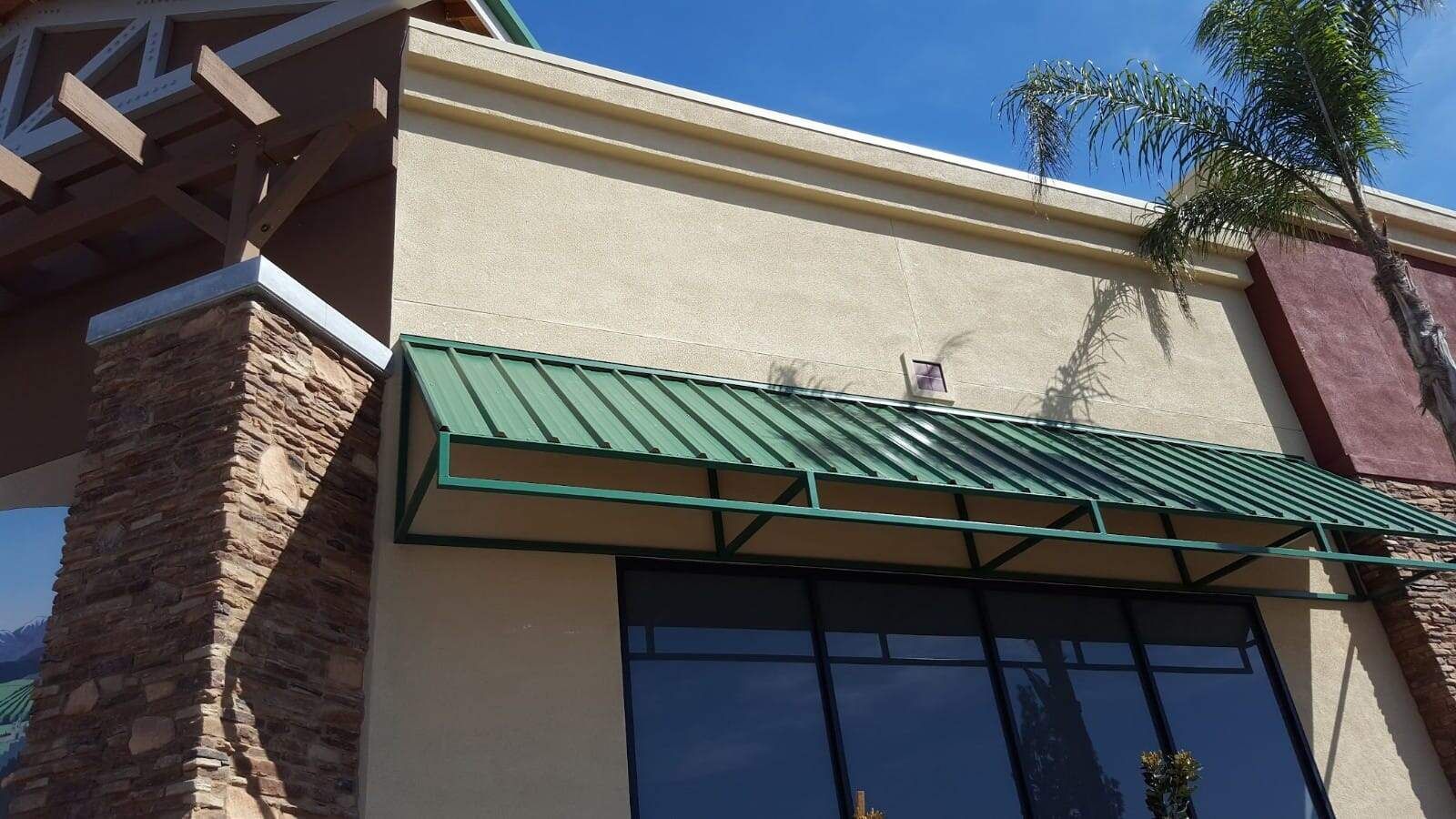 A cream-colored building exterior with a green metal awning above glass doors, next to a stone-clad pillar.