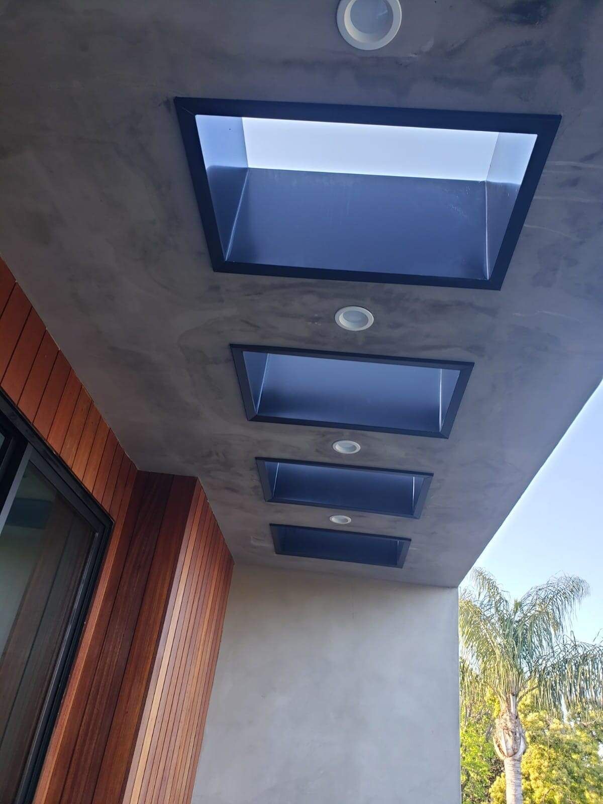 A gray concrete patio ceiling featuring four recessed rectangular skylights and small circular lights, beside a wood wall.