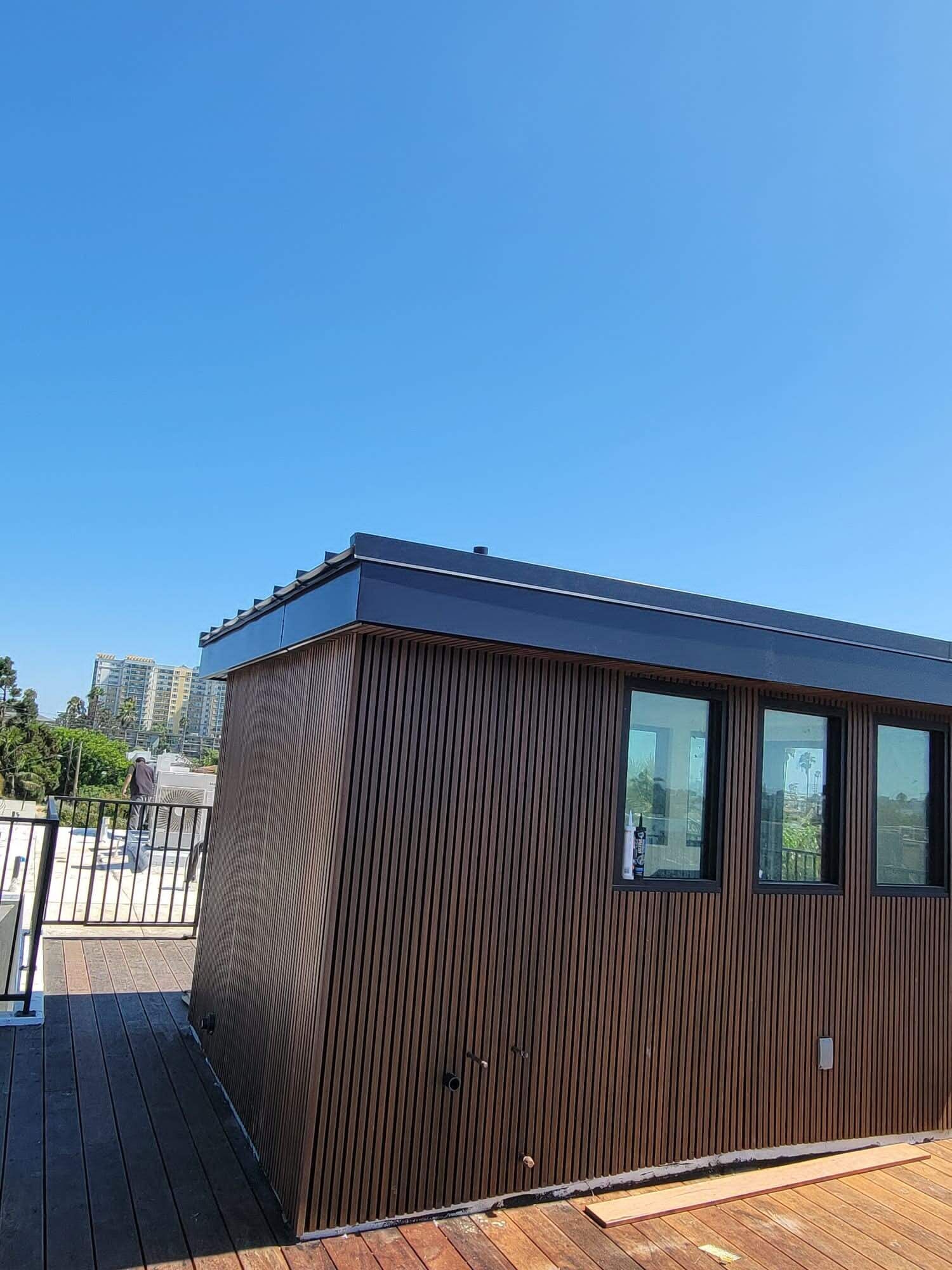 A modern wooden-slatted shed with three windows sits on a wooden deck against a clear blue sky.