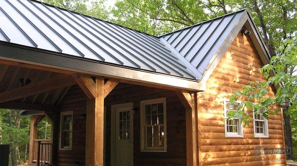 A small log cabin with a dark metal roof, featuring a covered porch with wooden support beams in a forest setting.