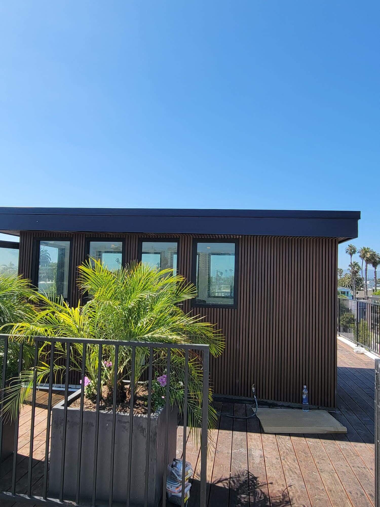 A small modern structure with vertical wood slats and glass windows sits on a paved patio beside a potted palm plant.