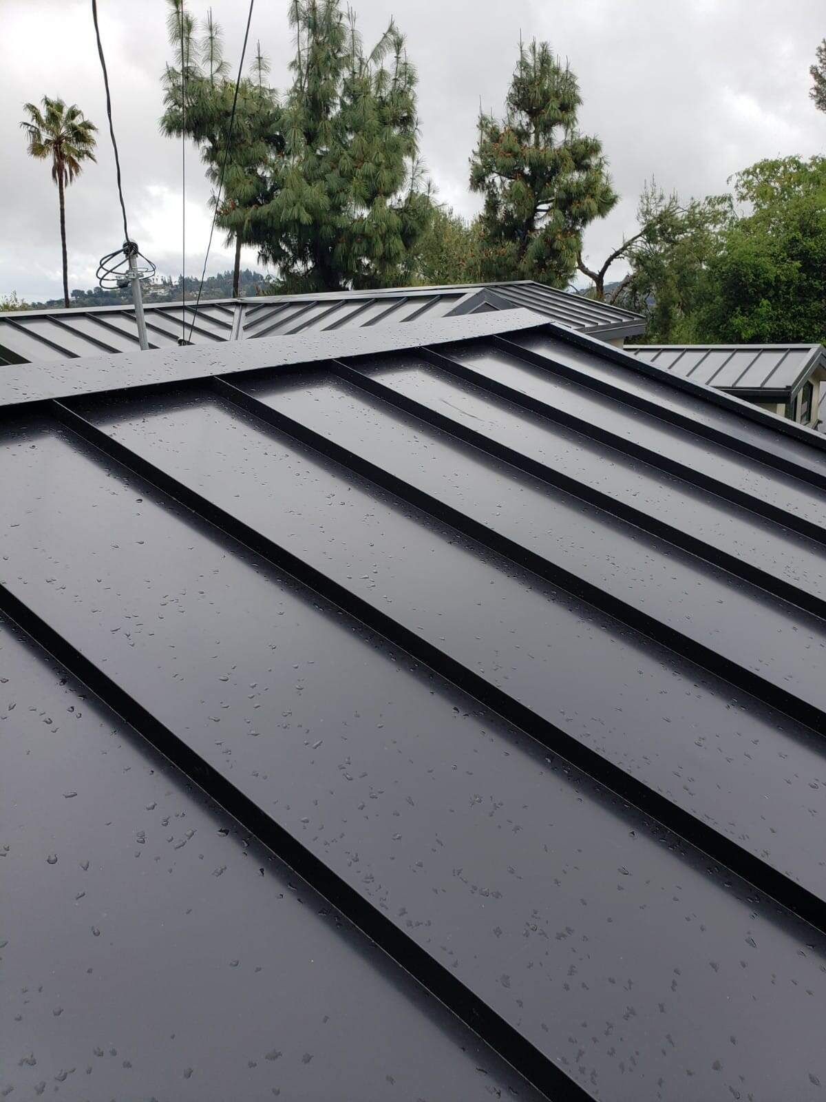 A close-up of a modern dark gray standing-seam metal roof with wet surfaces under a cloudy sky.