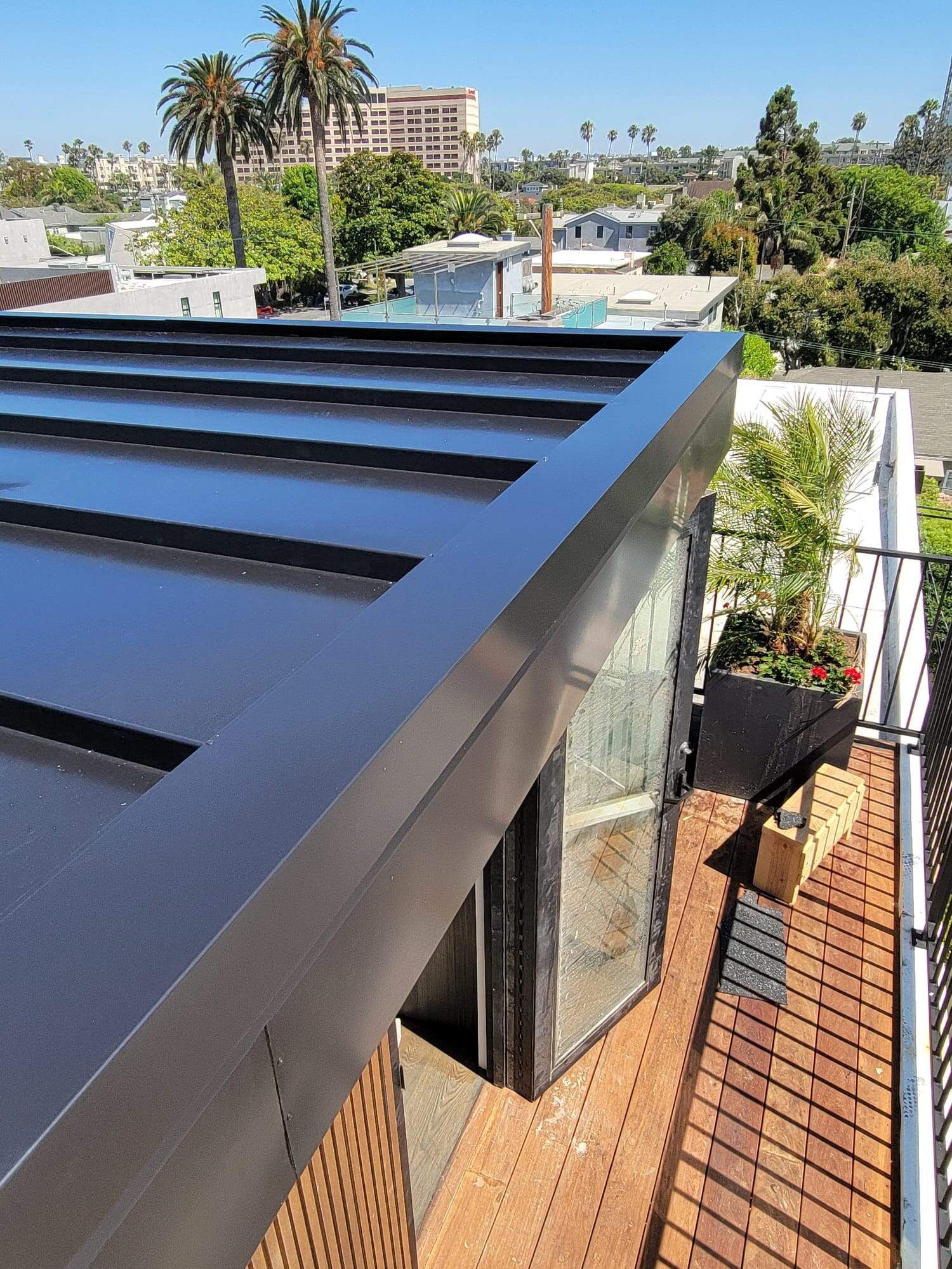 A high-angle view of a modern black metal roof overhanging a deck with brick flooring and a metal railing.