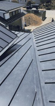 A high-angle view of gray metal roofing panels meeting at a central valley, with a house exterior and dirt pile behind.