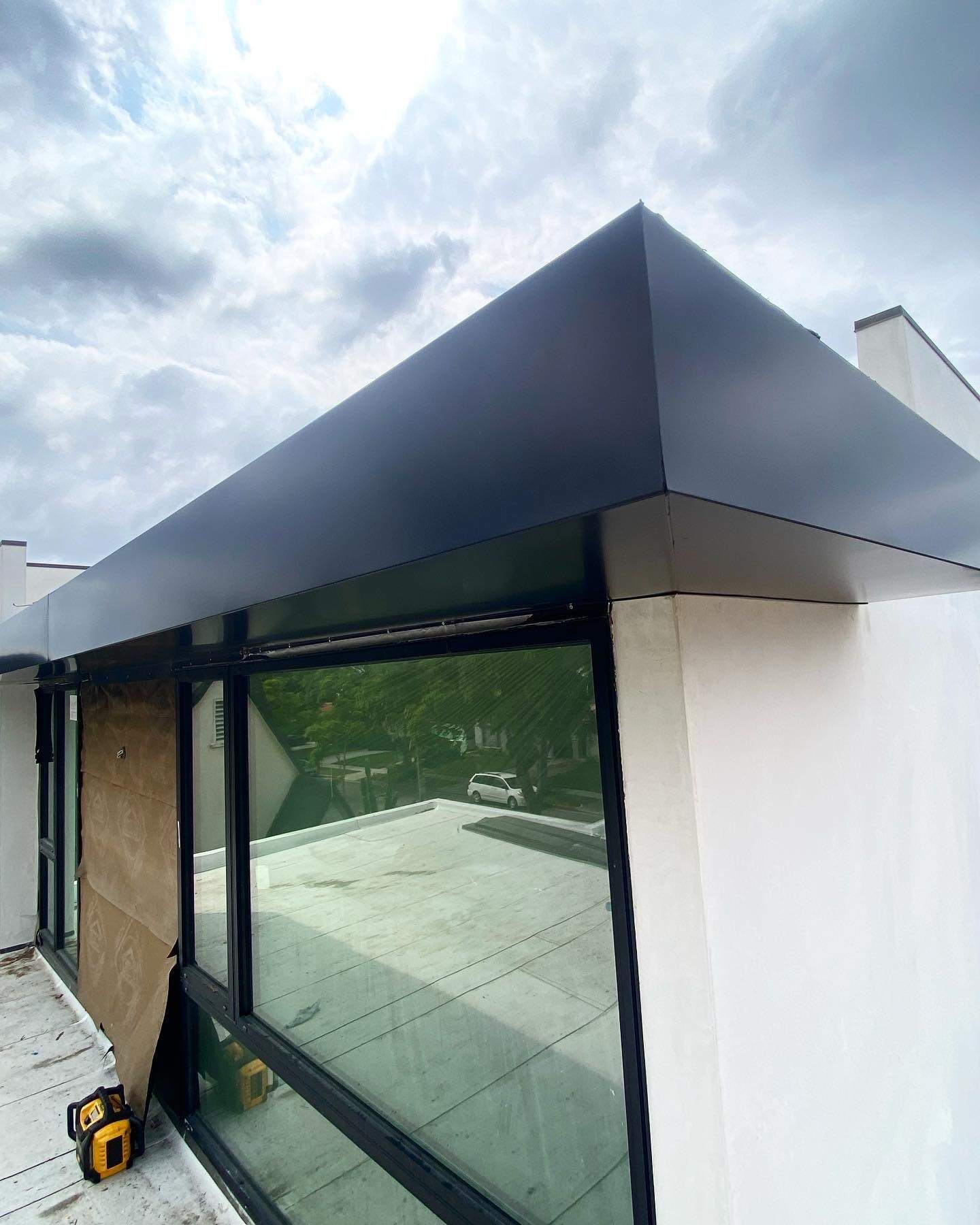 A matte black overhang covers a large window on a white exterior wall against a cloudy sky.