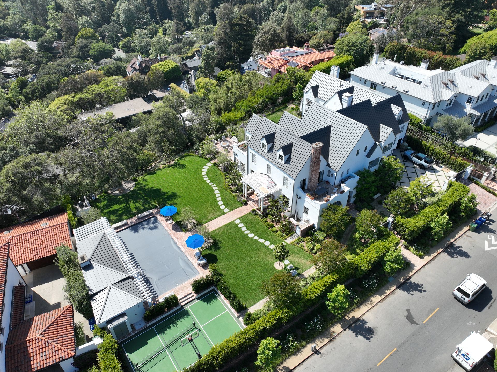 Aerial view of a large white house with a pool, tennis court, and surrounding trees on a sunny street.