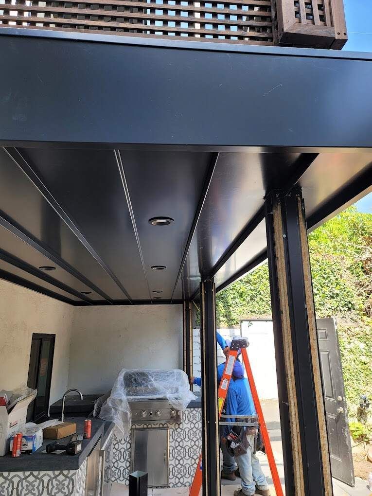 A person in a blue shirt works on an outdoor patio roof structure with black supports and installed recessed lighting.