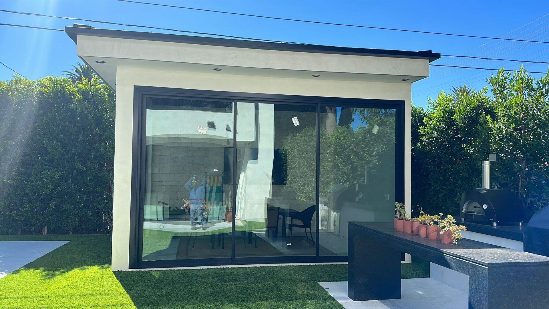 A small, modern shed with glass sliding doors stands in a sunlit backyard with green grass and lush hedges.