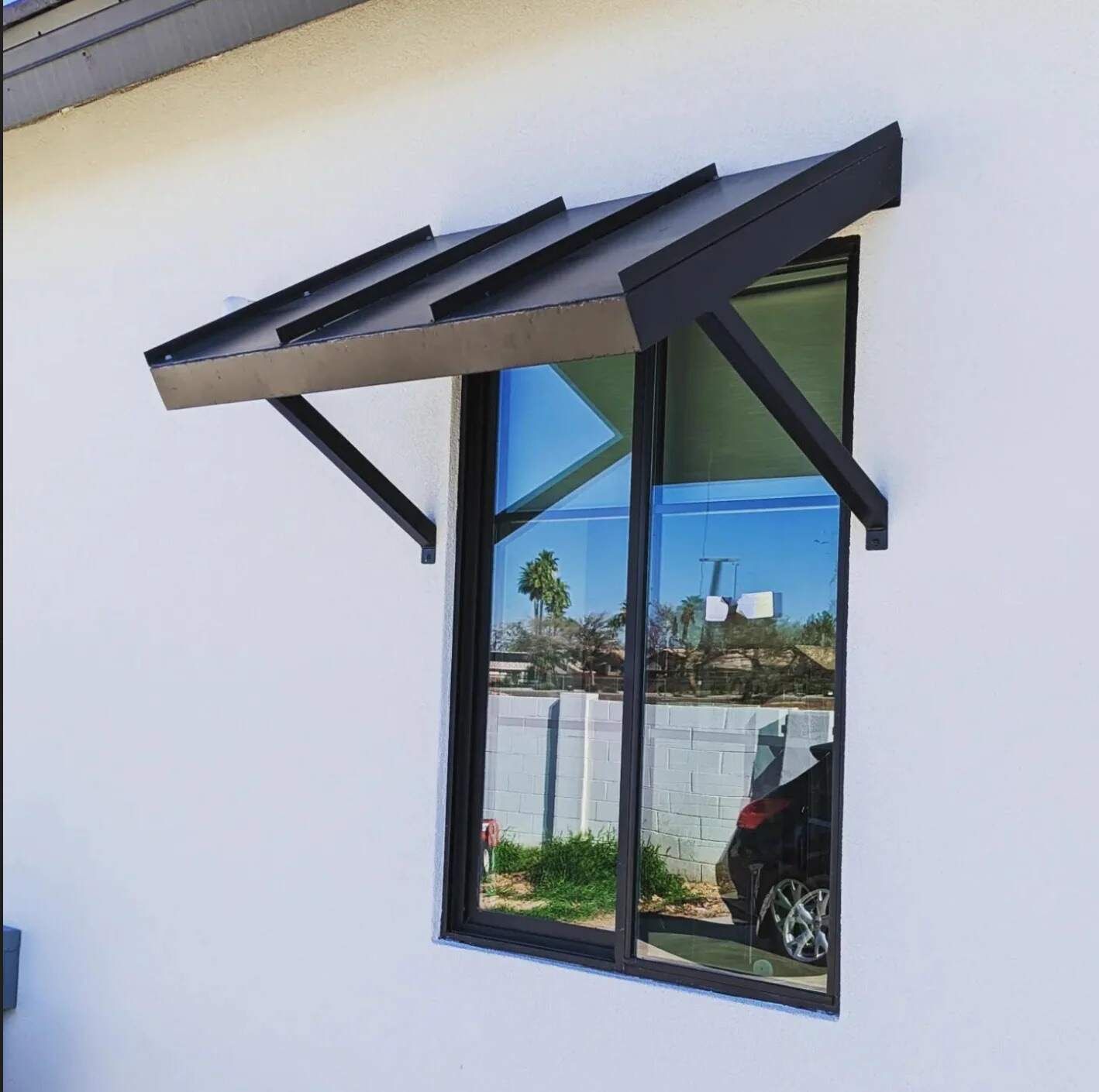 A modern, matte black metal awning mounted above a rectangular window on a smooth white exterior wall.