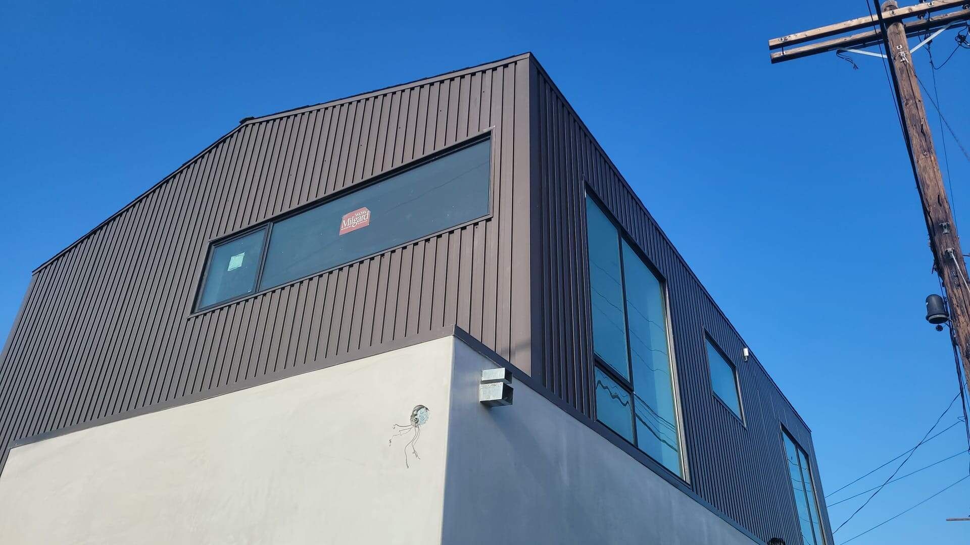 A modern, two-story building with a white stucco ground floor and a dark, textured metal upper level under a blue sky.