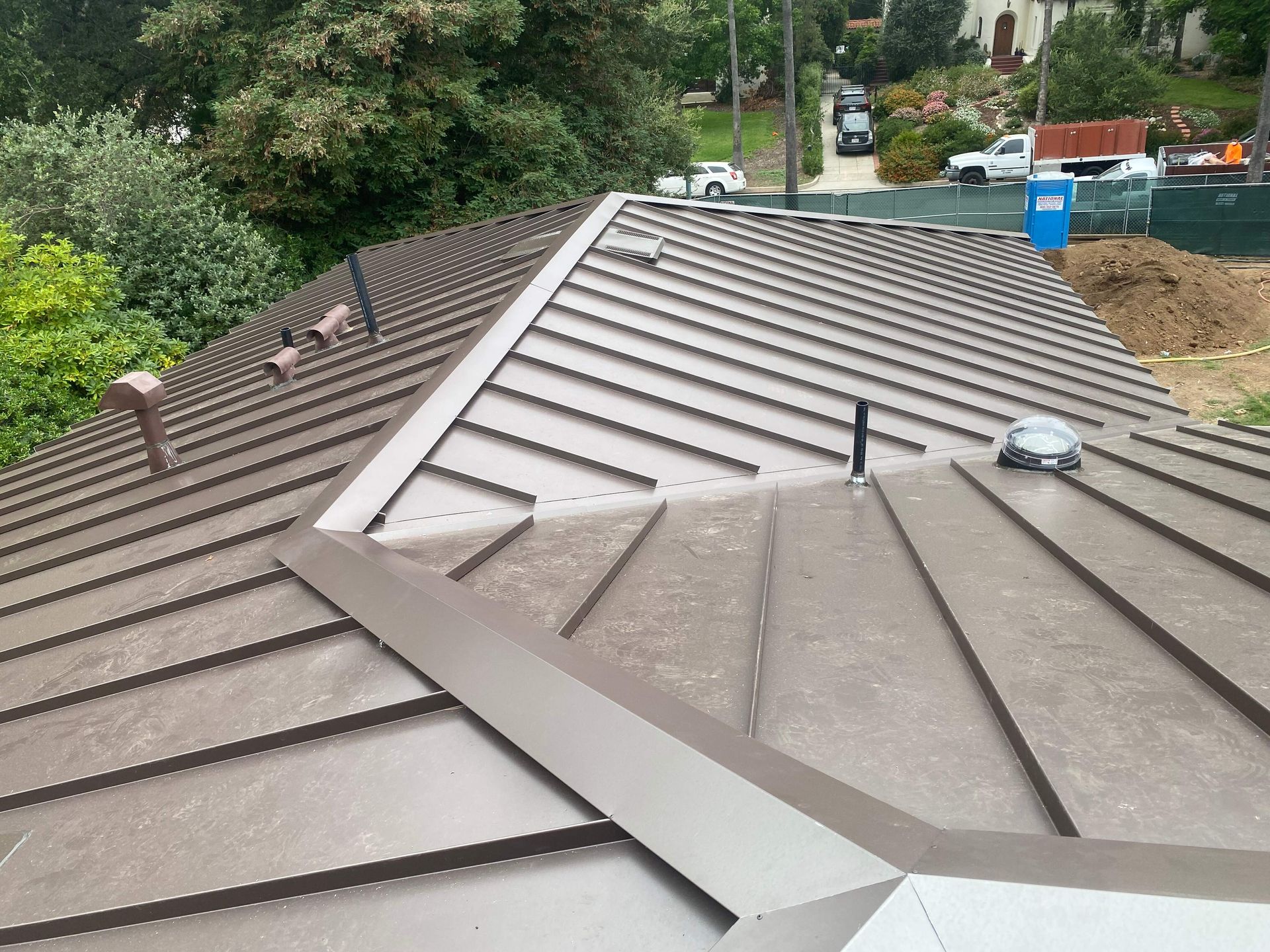 A high-angle view of a brown metal roof featuring raised seams, a central ridge, a pipe vent, and a circular skylight.