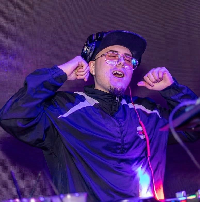 DJ in a cap and glasses dancing with headphones on under purple stage lighting at the Lady Hartford