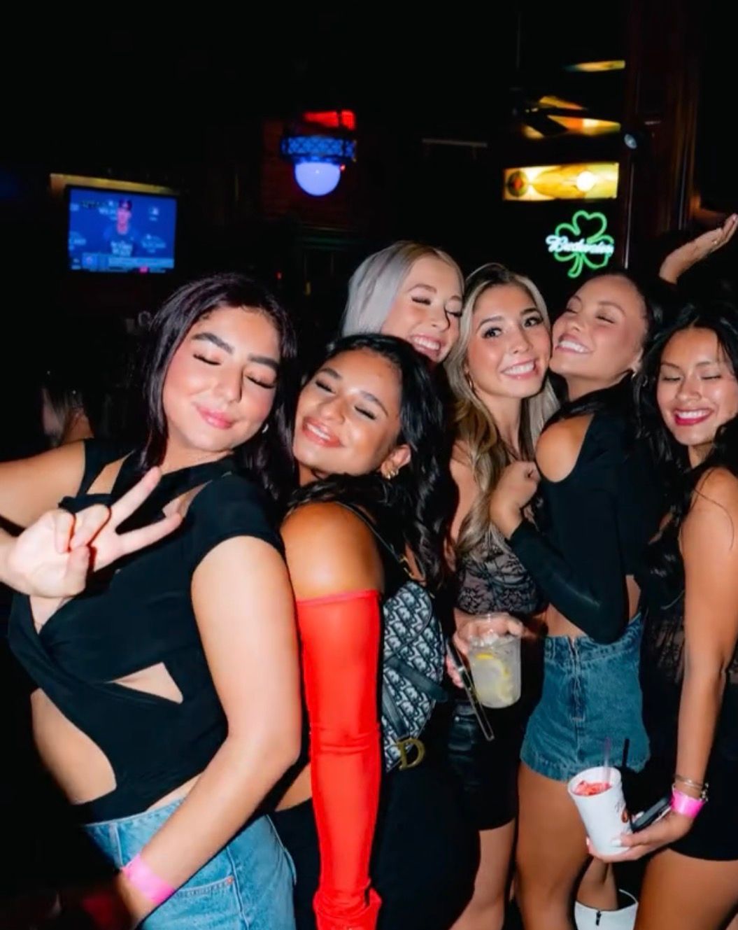 Five friends smiling and posing together in a dimly lit bar, one making a peace sign.