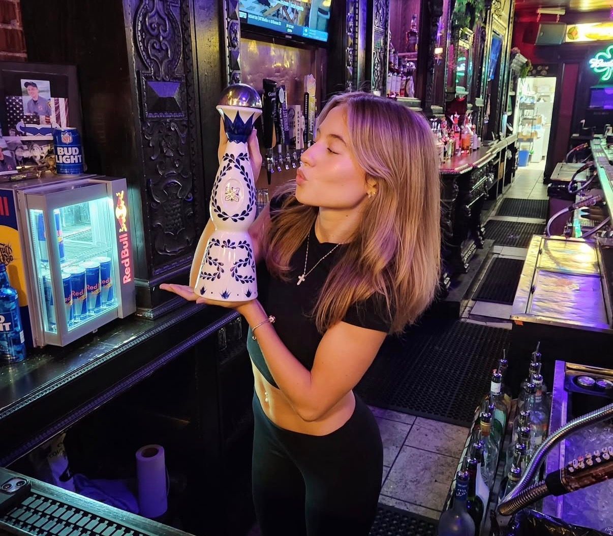 Woman in a neon-lit bar kissing a decorated bottle while holding it up.