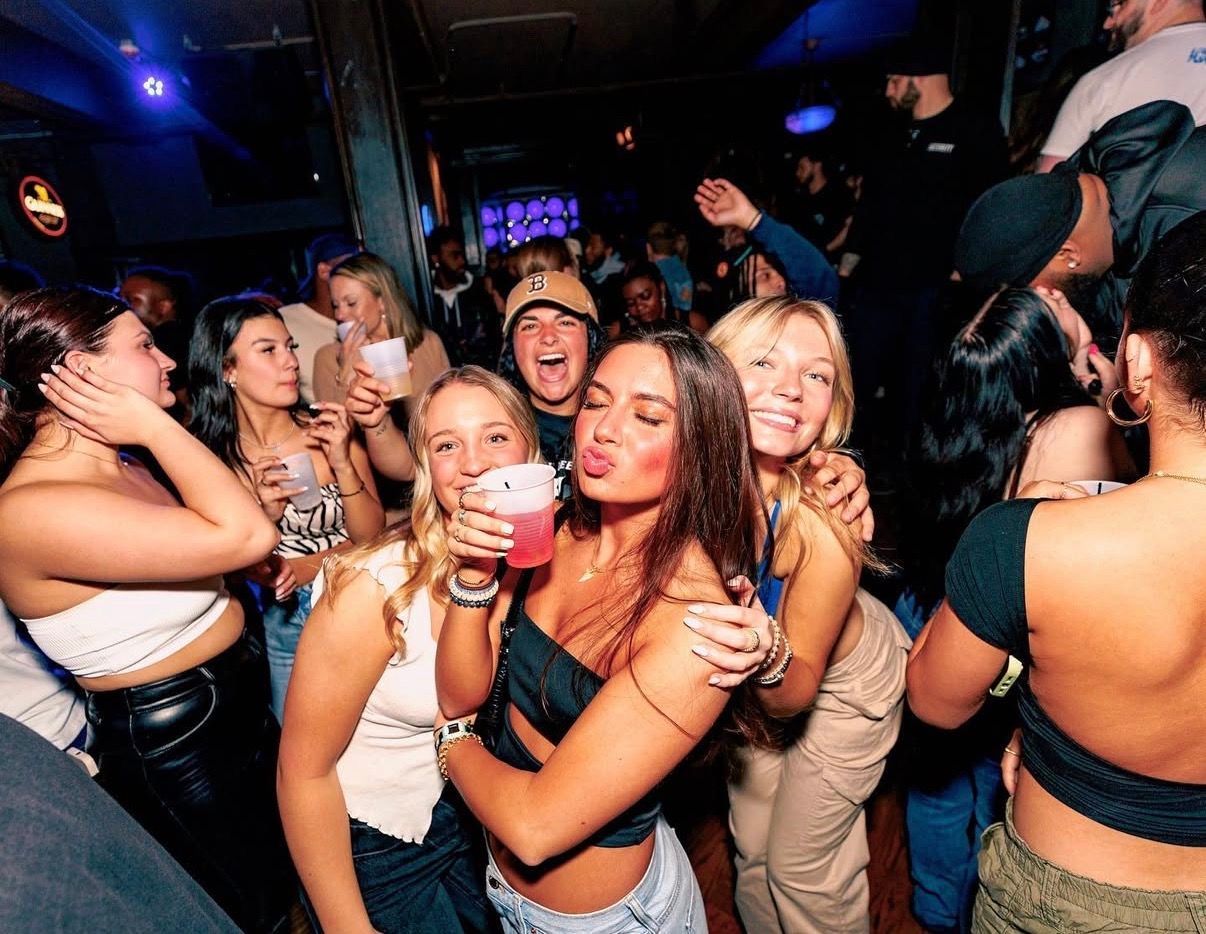 People dancing and posing with drinks in a crowded nightclub, smiling under colorful lights at the Lady Hartford