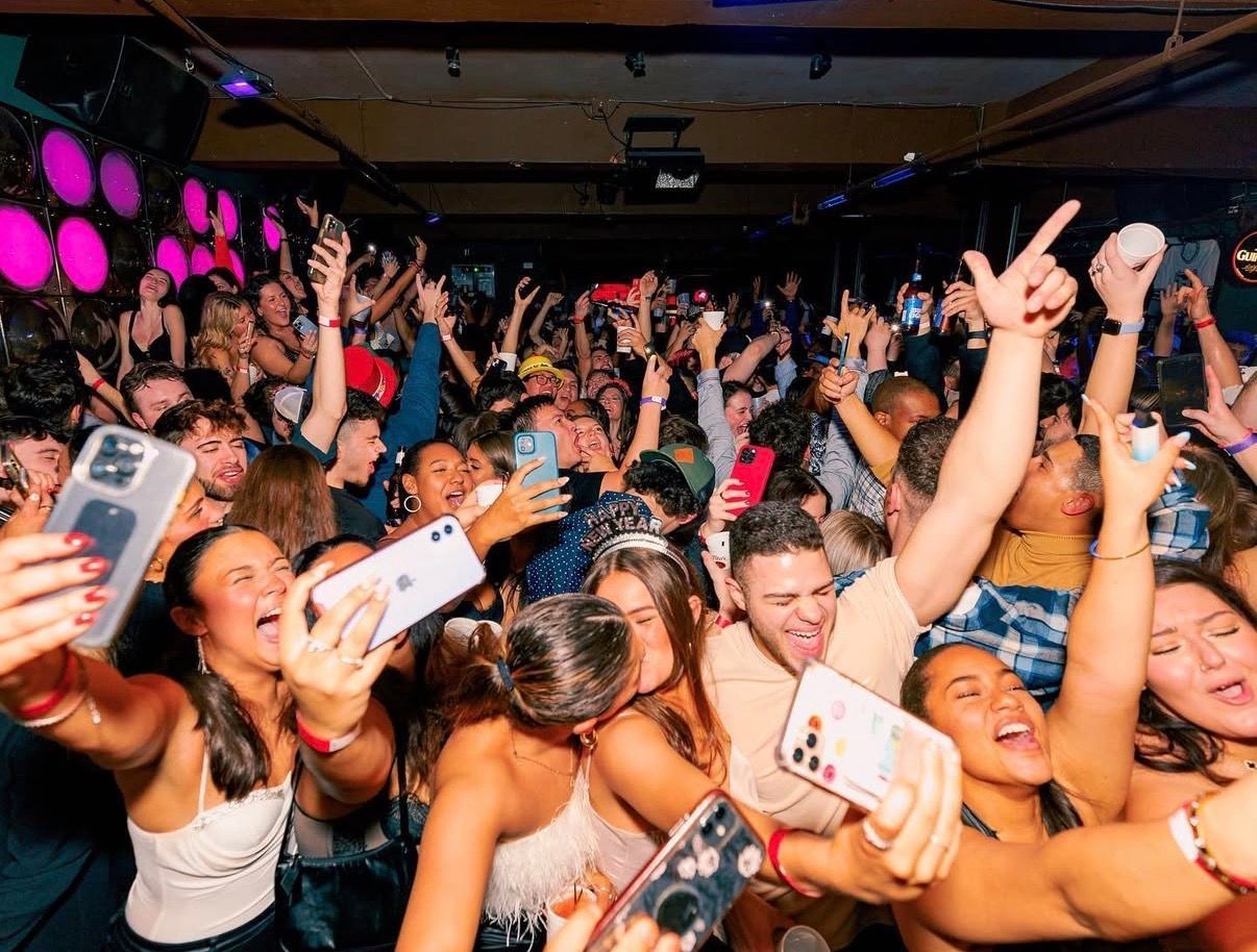 Crowded concert audience cheering with raised hands and phones under pink and purple lights at the Lady Hartford