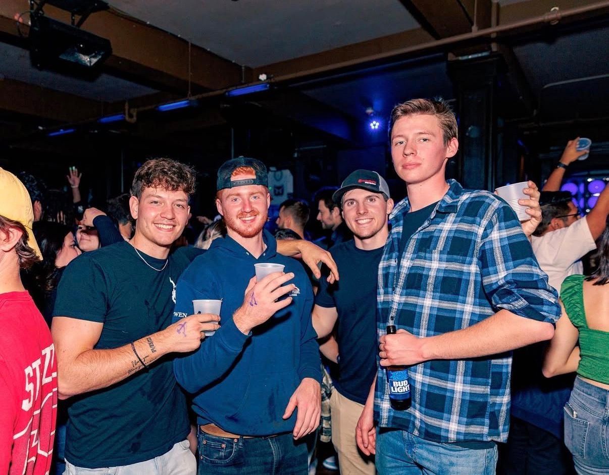 Four friends smiling and holding drinks at a crowded indoor party, one in a blue plaid shirt at the Lady Hartford