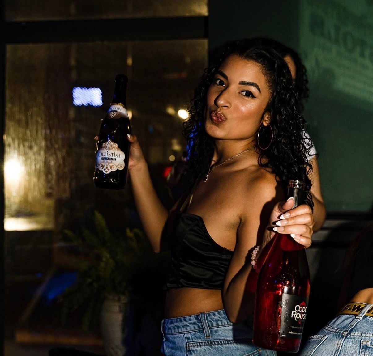 Woman in a dark bar holding two beer bottles, wearing a black top and jeans, with a drink on the table at the Lady Hartford