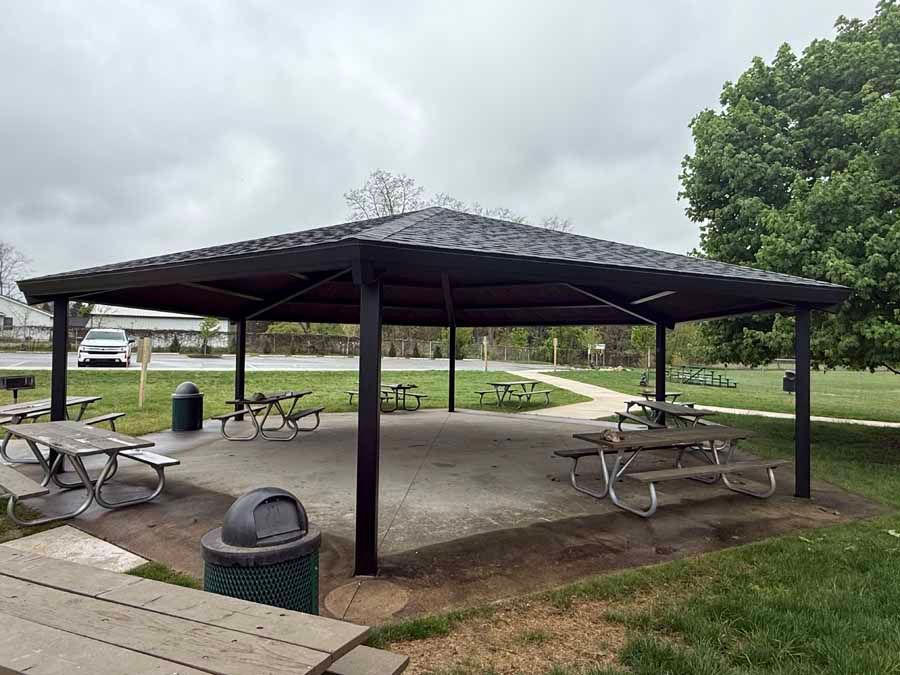Park pavilion with picnic tables, benches, and trash cans in a grassy area under cloudy skies