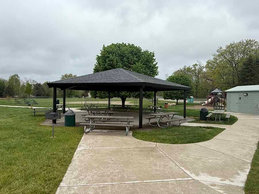 Park pavilion with picnic tables beside a paved path in a grassy park under cloudy skies