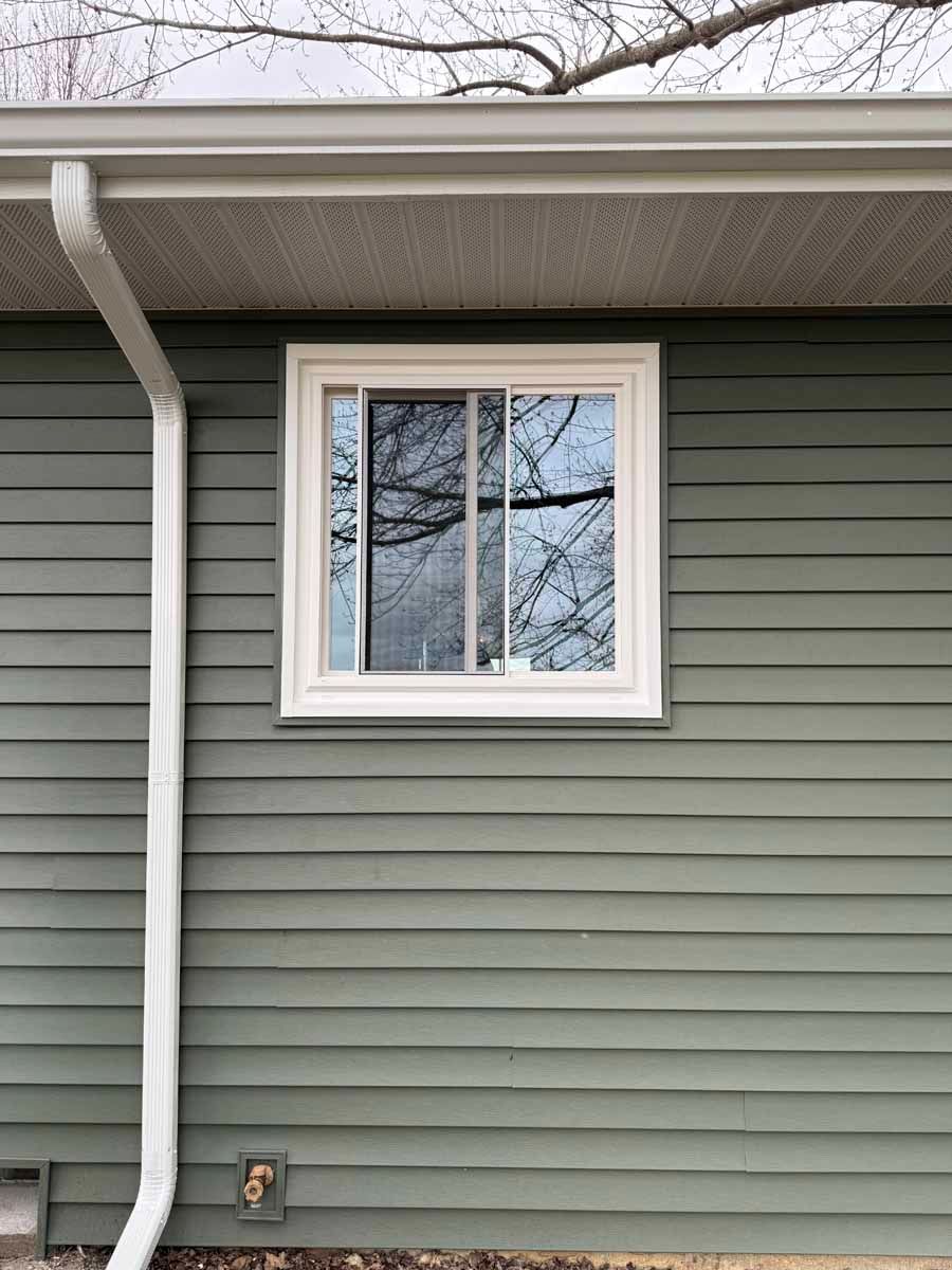 House exterior with gray siding, white-trimmed window, gutter, and downspout under the roof eave