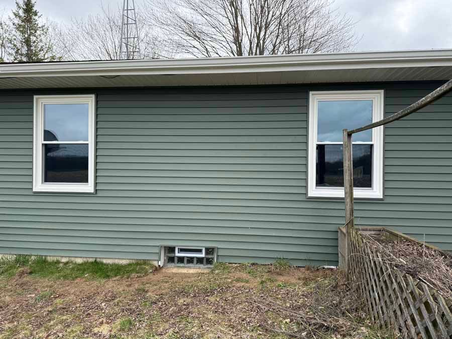 Side of a gray house with two windows, a small basement vent, and a fenced yard under bare trees.