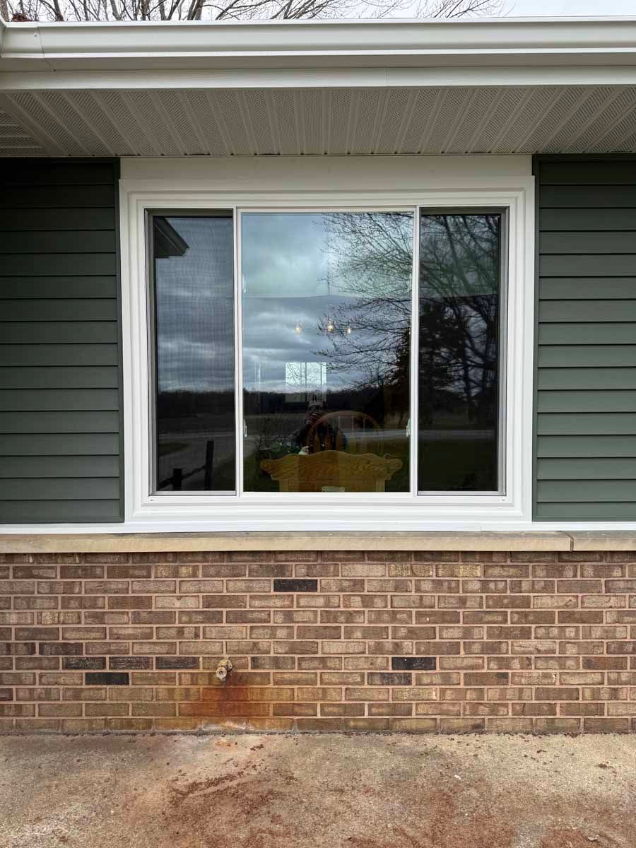 Window on green siding above brick wall, with rust-colored stain on the brick below.